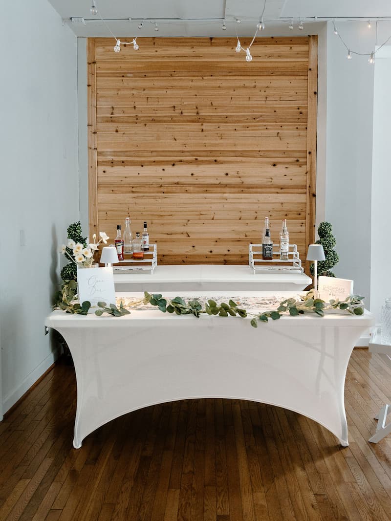 Classic DIY wedding bar setup with white spandex linen, eucalyptus garland and bar lamps