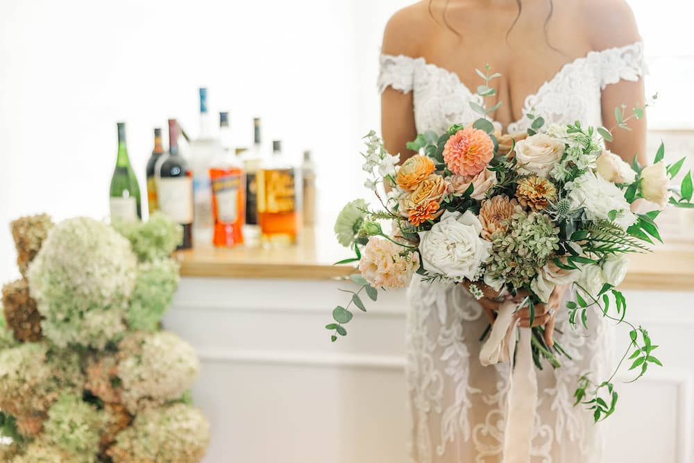 Bride in wedding gown holding bouquet in front of a styled wedding bar with full spirits display