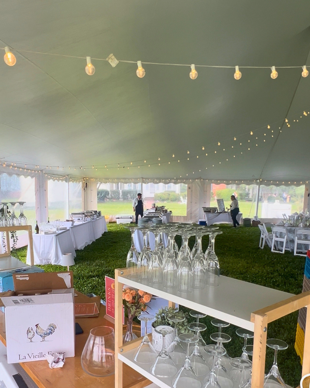 Stepne Manor tent interior during setup — glassware staged on Baker's Racks with bistro lights above